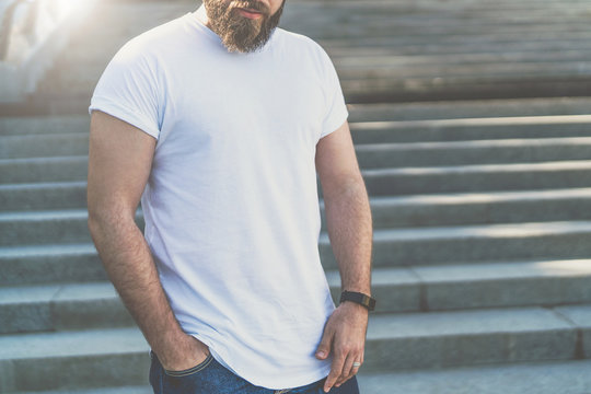 Front view.Young bearded hipster man dressed in white t-shirt is standing on steps with his hand in his jeans pocket. Mock up.