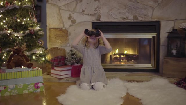 Cute Little Girl Sits By Cozy Fire And Christmas Tree, She Uses Binoculars To Search The Night Sky For Santa