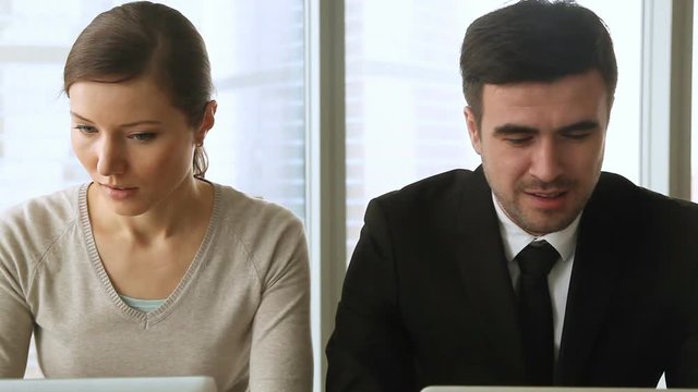 Young male and female rivals working together at office desk using laptops looking at each other, colleagues expressing instant dislike, hate or envy, coworkers showing jealousy of other&rsquo;s success
