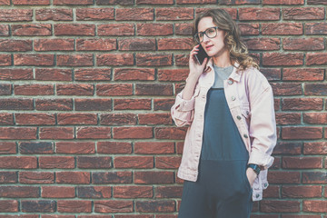 Young attractive hipster woman in glasses stands outside on a red brick wall background and talks on her cell phone.