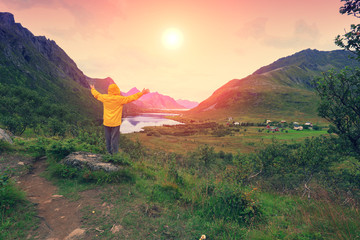 Panoramic view of the fjord. Twilight time with an amazing orange sky. The young man with hands in the air standing on a cliff of rock and gazing sunset. Beautiful mountain landscape at sunset.