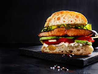 Ultimate sandwich with grilled salmon, cream cheese, sliced avocado and seasonals on black wooden board with free text space.