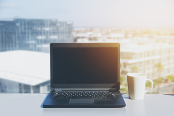 Fototapeta premium Blank screen laptop and cup on desk with view of city down below