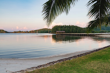 Sunset on the calm Thai beach. Tropical beach panorama with the smooth surface of the sea, palm trees, pier for boats and mountains in the distance, Koh Chang,Thailand.