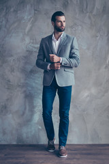 Full size portrait of stylish young bearded man standing on gray background. He looks stunning! in a suit, fixing the cufflinks