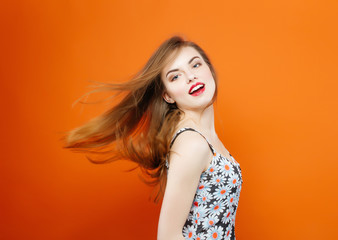 Active Young Woman with Long Dark Hair Wearing Dress with Flowers is Having Fun in Studio While Posing on Orange Background. Hair Style Concept.