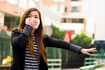 Young woman holding his protective mask with her hand on the street in the city with air pollution, asking for a taxi, city background