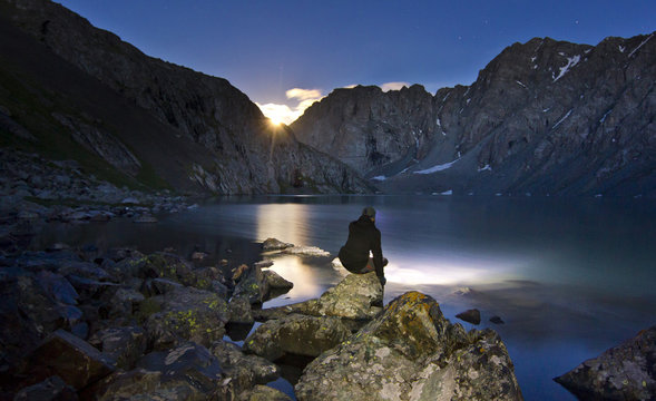 man in black jacket sitting near blue lake in mountains at night