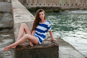 The girl with long hair in striped t-shirt hugging her knees sitting on a long stone pier. Early morning.