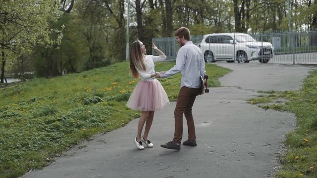 Young couple in love walking in summer park holding hands. Woman in pink tutu skirt man with longboard in hands Three-axis Gimbal stabilized tracking shot