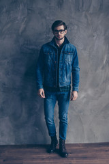 Autumn. Young serious bearded guy in casual jeans outfit is standing on a concrete wall`s background, wearing glasses and look pensive