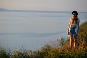 Young pretty woman wearing hat and sunglasses dressed in boho style clothes walking on the nature near water during beautiful sunset