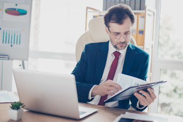 Mature business man is reading his notes, preparing for the meeting. He is broker, wearing suit and red tie, glasses, sitting in the modern office