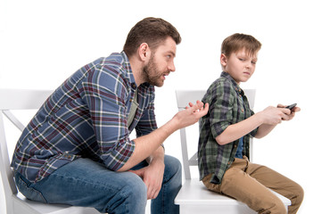 Fototapeta premium Father talking with little son sitting on chair and using smartphone, family problems concept