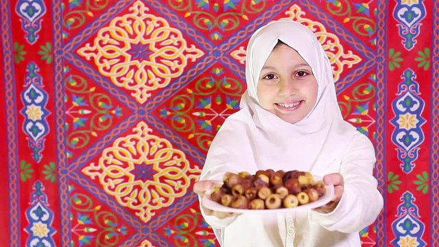 Little Muslim girl presenting a dish of dates for iftar - breaking fast in Holy Ramadan in front of islamic fabric background - showing generosity of Ramadan