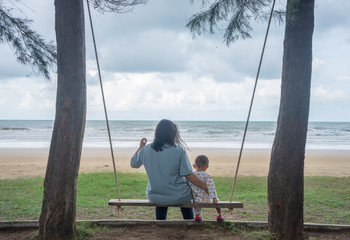 Mother and son are sitting on a swing at the beach, looking at cloudy sky and sea. Concept for...