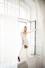Beautiful Blonde Woman Fiancee with Bridal Hairstyle, Event Makeup and Flowers Opening the Window