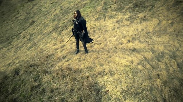 Bearded man dressed in assassin's costume is standing on the ruined fortress and drone is flying around him. He has sword in his hand. Top view.