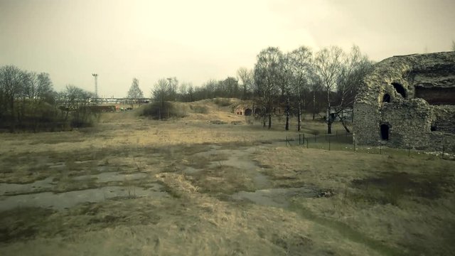 Bearded man dressed in assassin's costume is running along the field with sword in his hand. Ruffled hair. Top view from drone. Ruined fortress on background.