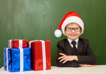 Happy boy in red christmas hat with gift boxes near empty green blackboard