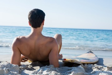 Rear view of shirtless man reclining by surfboard at beach