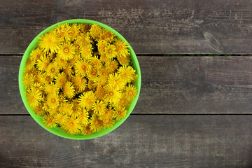 Green basket with yellow flowers of dandelions on the brown wooden background.