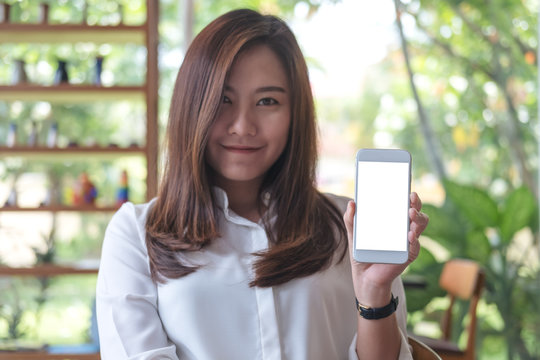 Mockup image of a beautiful woman holding and showing white mobile phone with blank white screen and smiley face in cafe