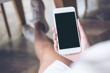 Mockup image of woman's hand holding white mobile phone with blank screen on thigh with wooden floor background in modern