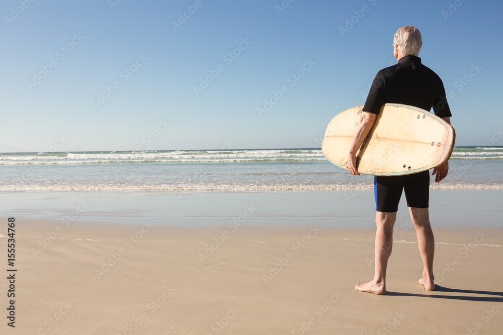 Wall mural Senior man with surfboard standing on sand  - Wall murals