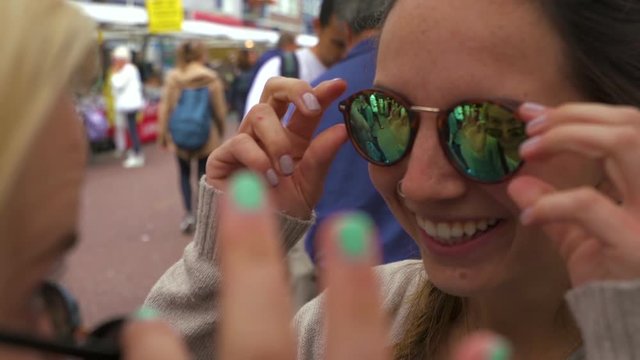 Young Women Try On Reflective Sunglasses And Look At Each Other, Girl Takes Her Pair Off And Smiles Wide