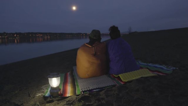 Couple Sit And Talk, Wrapped In Sleeping Bags, Enjoy View Of Moon Over Riverbank