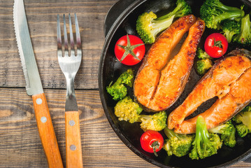 Healthy eating, diet. Baked grilled trout (salmon) with vegetable garnish - broccoli, tomatoes. In a portioned frying pan, on a wooden table. Top view copy space