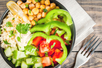 Healthy eating, diet. Vegetarian vegetable buddha bowl - couscous (quinoa), chickpeas, tomatoes, zucchini (cucumber), pepper, hummus, lemon and greens. On wooden table, with water, close top view