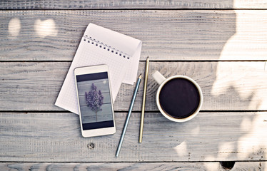 Cup of coffee, notepad, phone with photo of lilac branch on a white wooden table