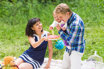 Fototapeta premium Happy young mother and father with their baby daughter relaxing on a blanket in a park celebrating with birthday cake