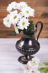 Close-up of flowers in iron vase on wooden table. Daisies and lilies. Vertical shoot.