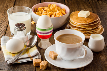 Breakfast table with coffee, boiled eggs, pancakes, cornflakes and milk.