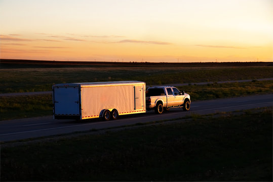 A white pickup truck pulls a small white box trailer down a rural highway during sunset hours. All visible markings and trademarks have been removed.