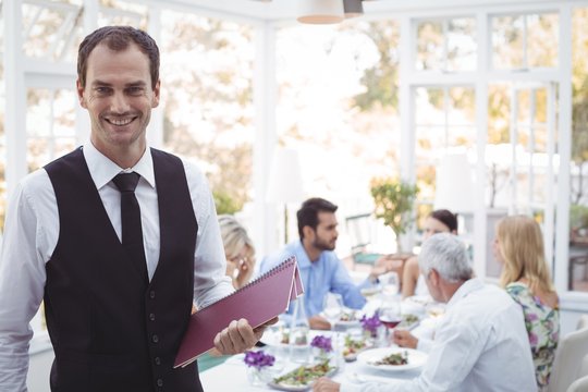 Smiling waiter holding menu while friends dining in background