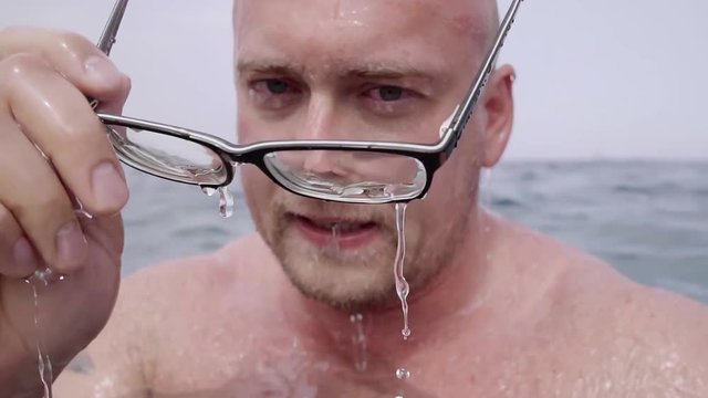 Successful young unshaven and bald man emerges from under the water of the sea wears glasses and shows thumbs up. Close-up.