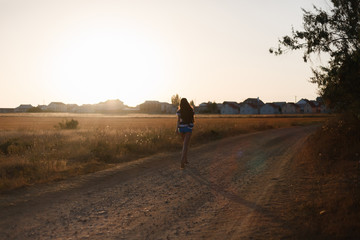 The girl with long hair running and enjoys raising his hands up on the sand road at sunset along the field with dry grass