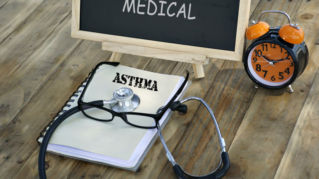notebook with the words "ASTHMA" and stethoscope, glasses, chalk board, alarm clock on a wooden table. medical and healthcare concept.