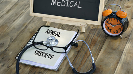 notebook with the words "ANNUAL CHECK-UP" and stethoscope, glasses, chalk board, alarm clock on a wooden table. medical and healthcare concept.