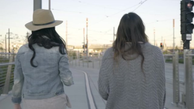 Two Young Women Walk (Away From Camera) In City, Commuter Train Passes By 