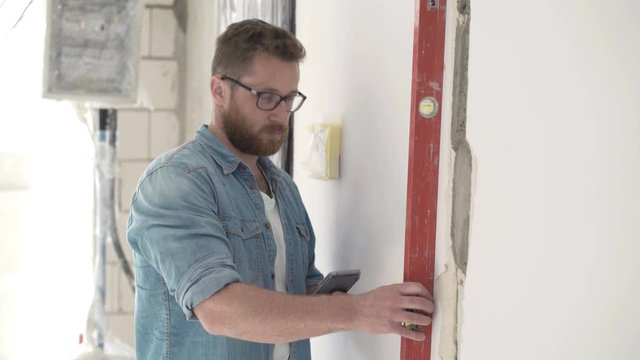Male architect with smartphone checking wall with level at new home 

