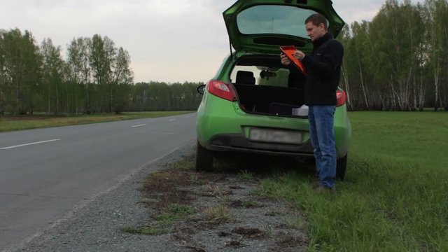 Young man with broken car on the rural road takes triangle from car trunk