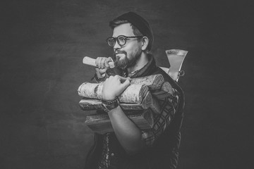 Bearded male in eyeglasses dressed in fleece shirt holds firewoods and axe on his shoulder.