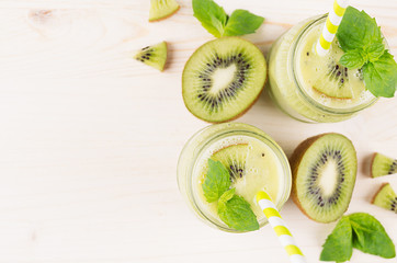 Fototapeta na wymiar Freshly blended green kiwi fruit smoothie in glass jars with straw, mint leaf, cute ripe berry, top view. White wooden board background, decorative border, copy space.
