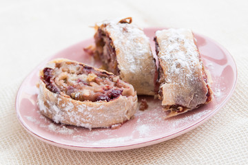 Apple strudel with icing sugar, cherries and raisins on rosa plate
