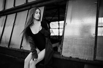 Sexy model girl wit long legs at black lingerie outfit body swimsuit combidress and jacket posed at the roof of abadoned industrial place with windows.